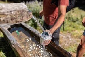 Ein Herr hält seine Wasserflasche zum Brunnen hin um etwas Wasser einzufüllen doch es plätschert alles über. | © Alpbachtal Tourismus | shootandstyle