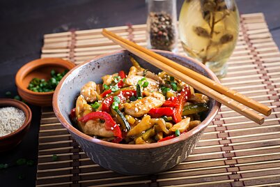 Eine Schüssel mit einem chinesischen Gericht steht auf einem Tisch mit Bambusunterlage, Stäbchen und einem Getränk. | © freepik