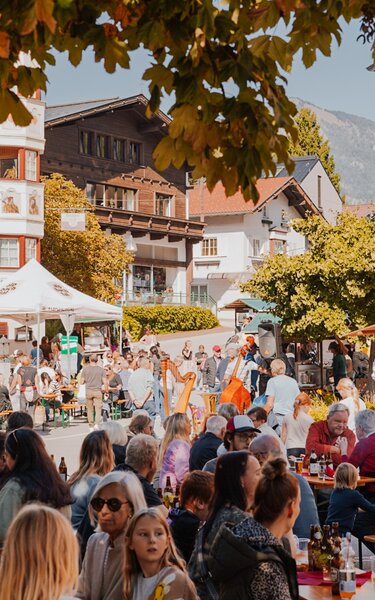 Weitwinkelbild über den Herrnhausplatz, aufgestellte Biertische mit zahlreichen flanierenden Personen, Martkständen und einer Musikgruppe mit Harfe, Kontrabass und Steirischer Harmonika. | © Brixlegger Wirtschaft | Genusskind