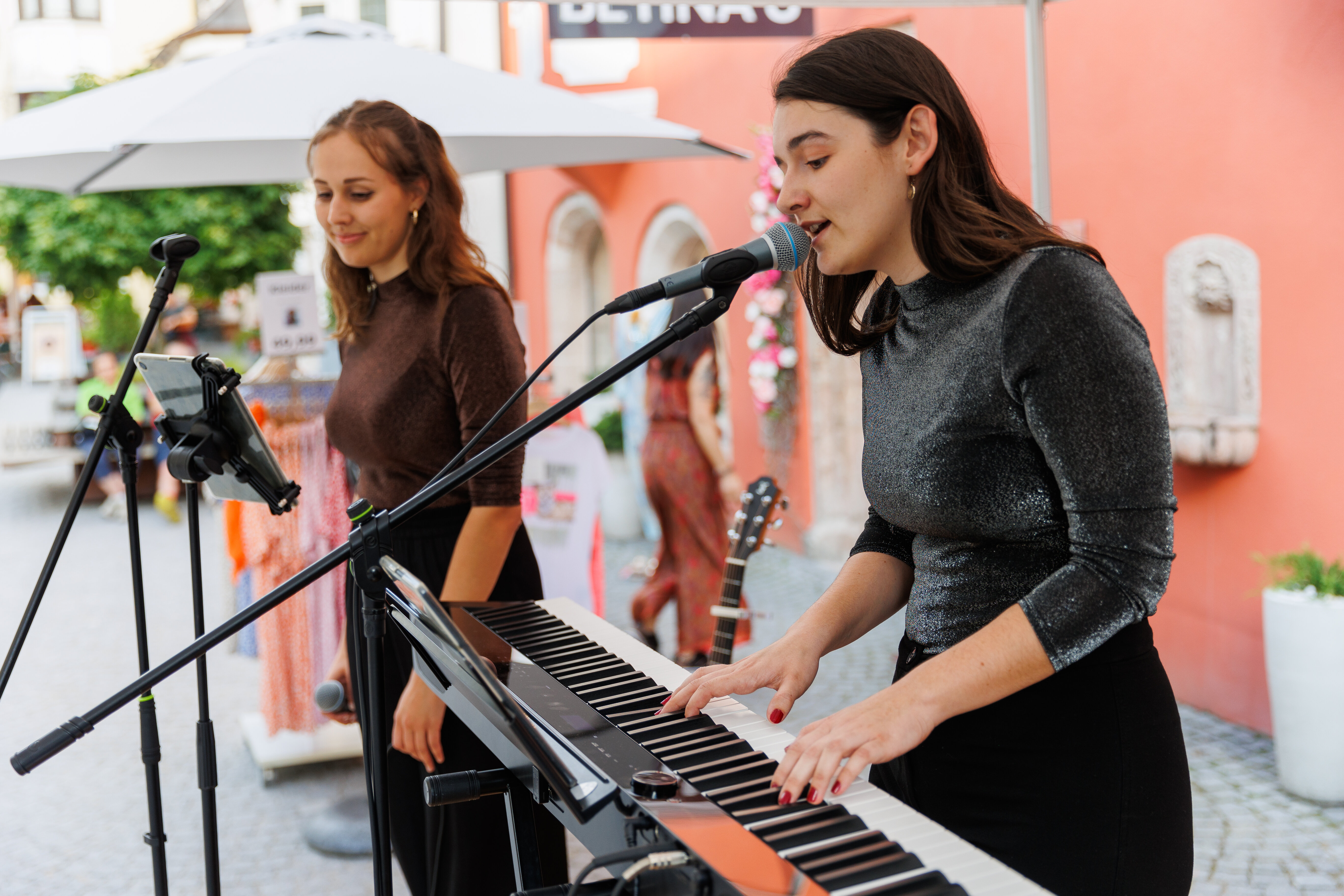 Zwei junge Frauen stehen vor Mikrofonen und einem Klavier, im Hintergrund steht eine Gitarre und Kleiderständer vom Geschäft dahinter. | © WiR Alpbachtal | Maximalgrafik