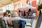 Stimmung beim Nightshopping Rattenberg | © WiR Alpbachtal | Maximalgrafik Personen sitzen auf der Gastterrasse eines Cafes in der Stadt Rattenberg, sie schauen auf eine kleine Bühne mit zwei Musikerinnen mit Klavier und Gitarre, welche unter einem Zelt mit der Aufschrift "Alpbachtal" geschützt sind. | © WiR Alpbachtal | Maximalgrafik