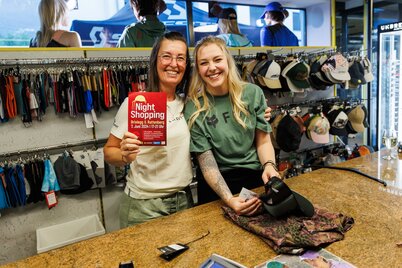 Zwei Verkäuferinnen von Connys Sport und Mode Brixlegg stehen hinter der Kassatheke und zeigen einen Flyer mit der Aufschrift "Night Shopping", im Hintergrund hängen Schildkappen und Mützen. | © WiR Alpbachtal | Maximalgrafik