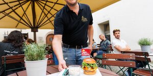 Night Shopping im Dun Aengus Rattenberg | © WiR Alpbachtal | Maximalgrafik Ein Mann bringt einen Burger mit einem kleinen Fähnchen mit der Aufschrift "Night Shopping" auf einem Teller garniert zu einem Tisch im Gastgarten des Dun Aengus Rattenberg. | © WiR Alpbachtal | Maximalgrafik