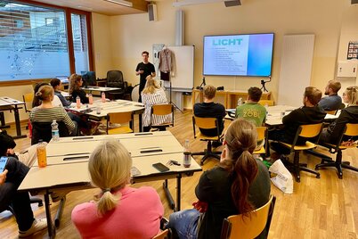 Personen sitzen in einer Schulklasse und hören einem Vortragenden zu, welcher zu einer Präsentation spricht, auf der elektronischen Tafel ist eine Folie mit der großen Überschrift "Licht" zu lesen.  | © WiR Alpbachtal