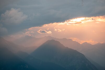 Blick vom Wiedersbergerhorn auf die Berge Richtung Stans, Wolken hängen herein, Sonnenstrahlen färben die Wolken orange. | © WiR Alpbachtal | Shoot&Style