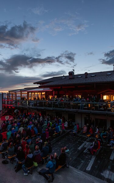 Menschen sitzen vor einer großen Kinoleinwand auf der Terrasse eines Bergrestaurants, die Stimmung ist abendlich. | © WiR Alpbachtal | Shoot&Style